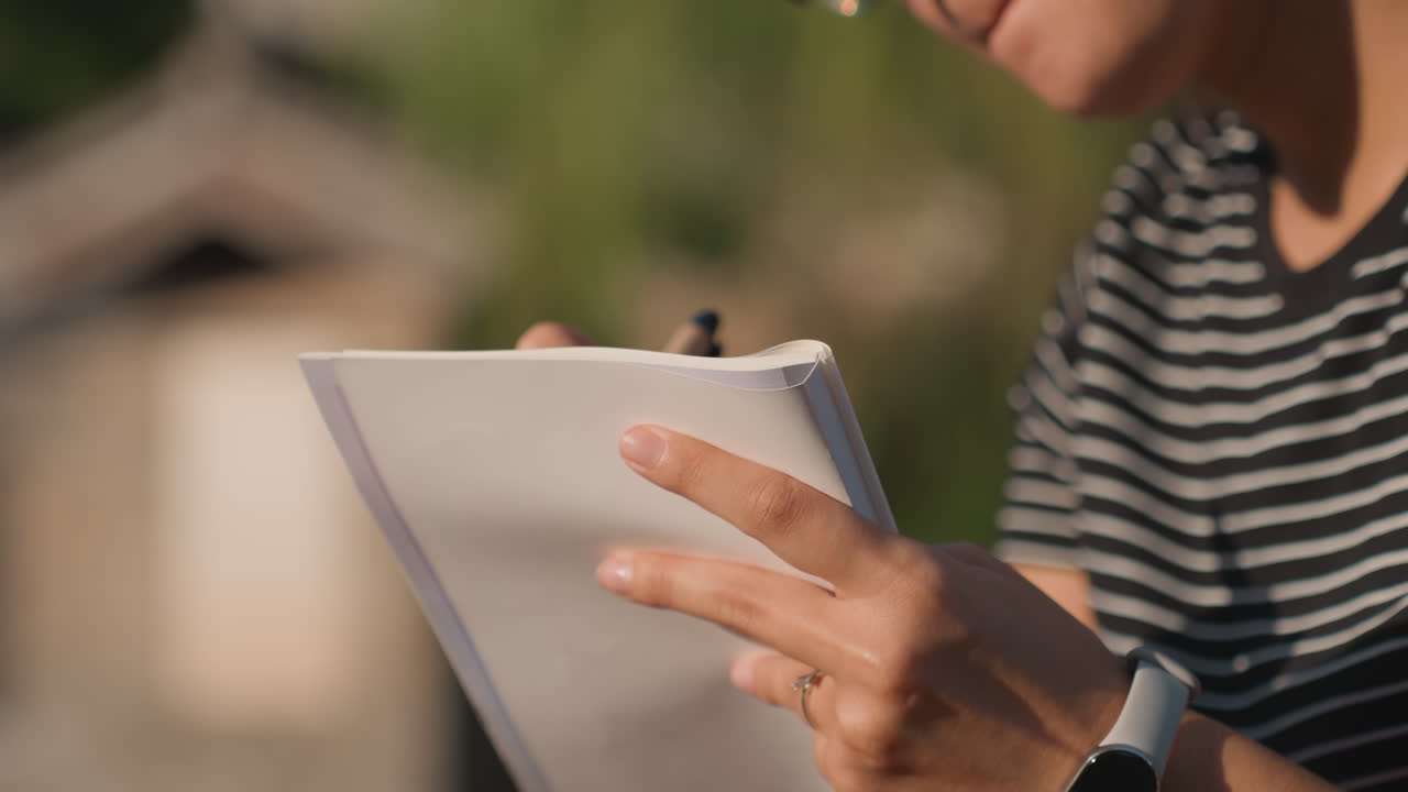 Donna bianca che scrive su un taccuino all'aperto, primo piano delle mani che tengono penna e carta, camicia a righe, smartwatch e anello, calda luce del tramonto, sfondo sfocato di tetti urbani, umore riflessivo e spontaneo.