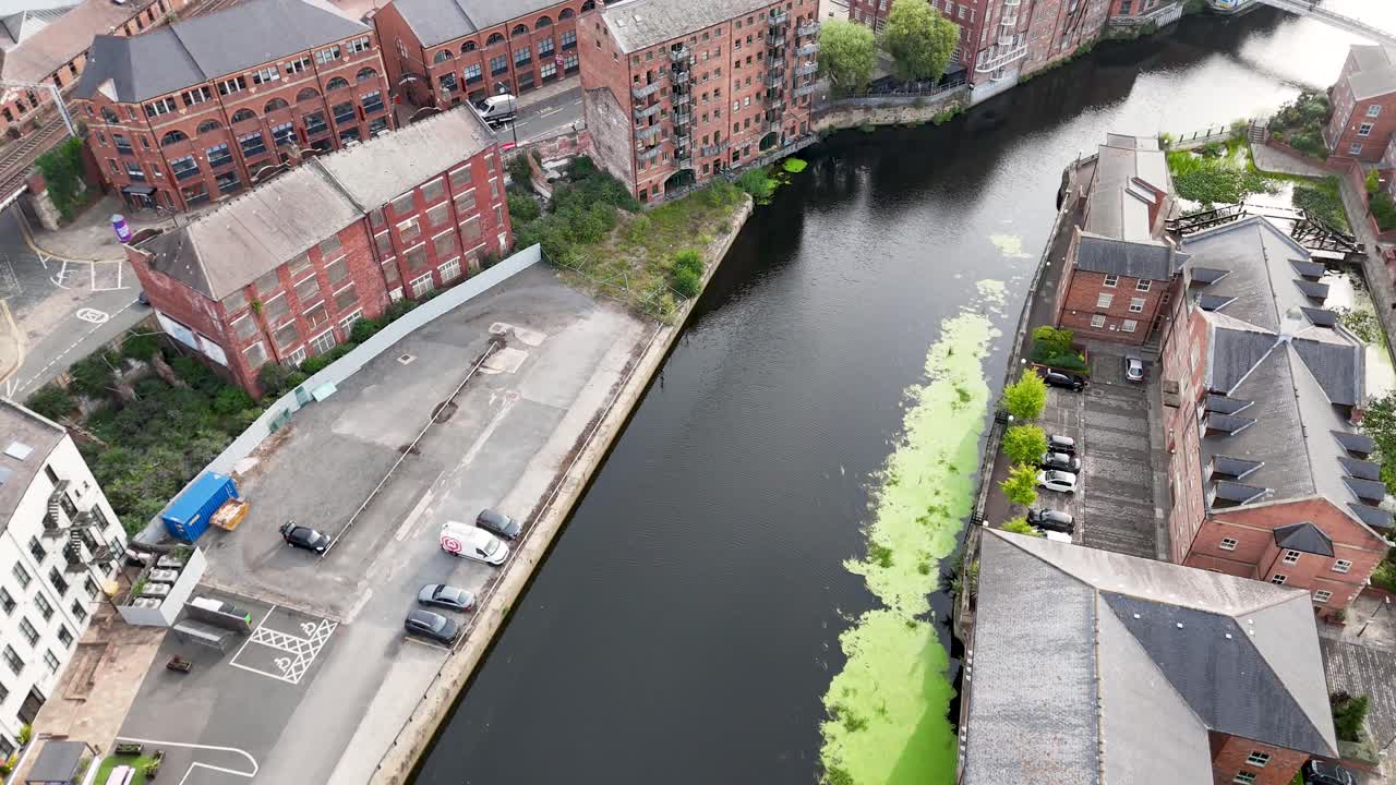 High-angle aerial footage glides above the River Aire, revealing historic brick warehouses, parked vehicles, and green riverbank vegetation under diffuse daylight