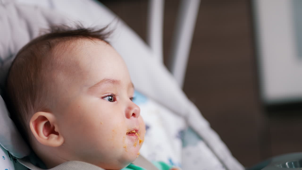 Active Caucasian child sitting in a baby chair being fed. Cute boy eats and waves his hands. Blurred backdrop.