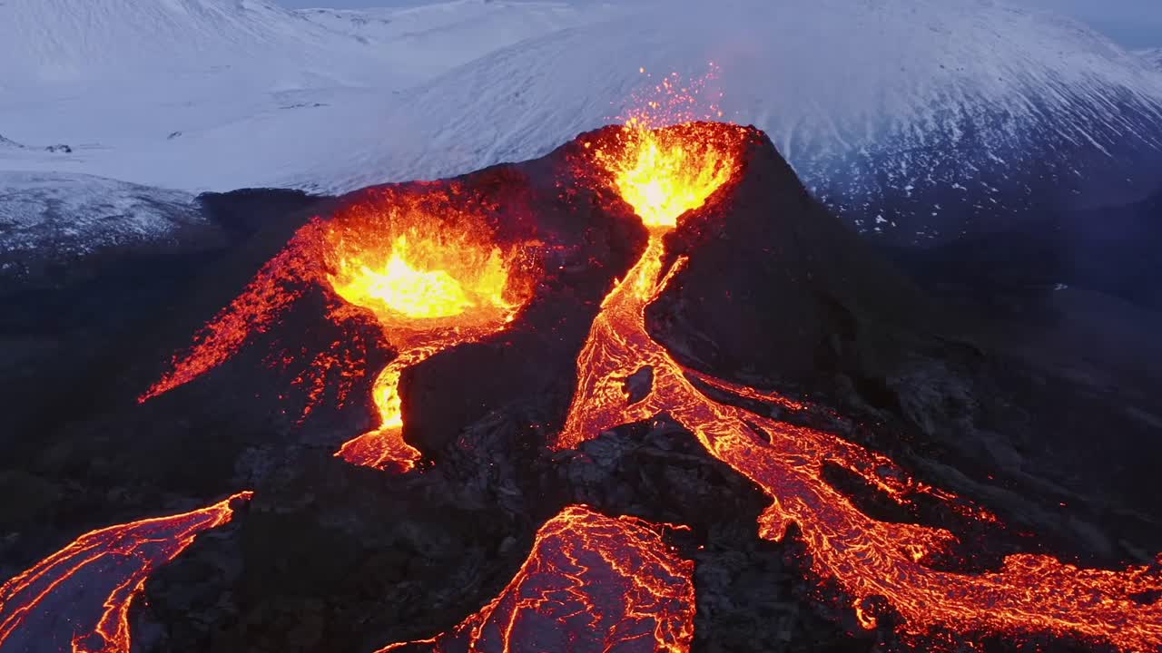 Aerial cinematic shots from a 4K drone capture two volcanic openings spewing lava, which then cascades down below, set against a backdrop of snowy wintry Alps