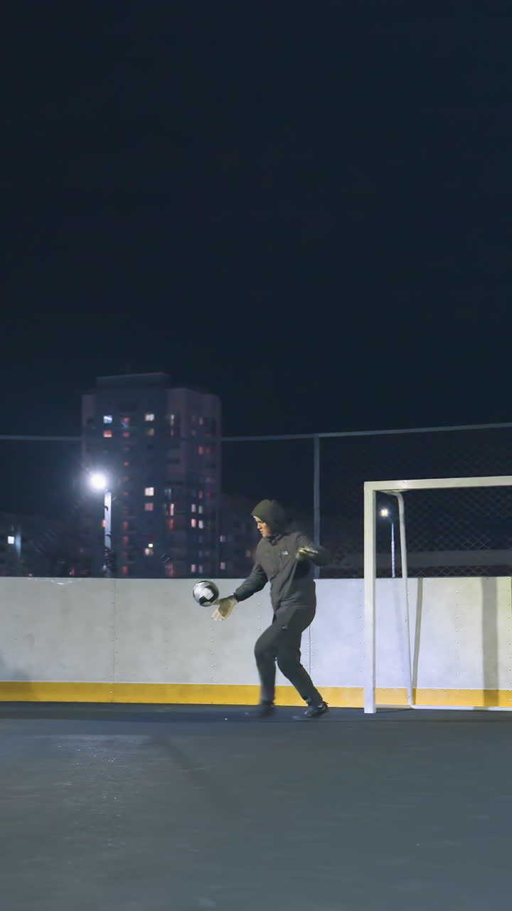 Footballer in dark sportswear kicks soccer ball out of the goal area on an urban field at night, surrounded by tall buildings, this dynamic action conveys determination under bright floodlights