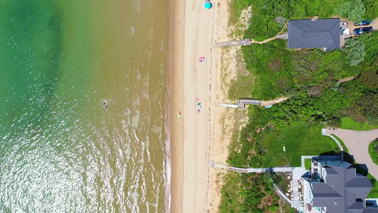Top-down drone video captures a pristine shoreline where beach houses overlook sandy stretches, stairways descend to the beach, and turquoise surf sparkles in bright sunlight