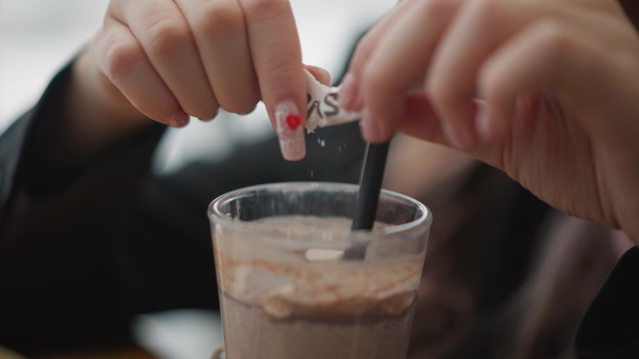Mani che spezzano una zolletta di zucchero sopra un caffè freddo mentre mescolano con un cucchiaino all'interno di un bicchiere, mostrando il movimento di dolcificazione, vortici cremosi, la consistenza di una bevanda fredda, luce soffusa interna, messa a fuoco selettiva e un caffè casual.