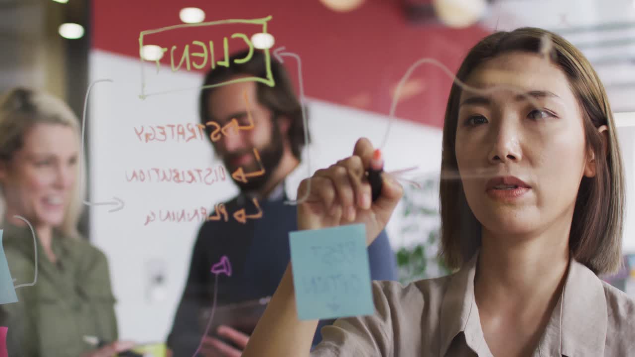 mujer asiática escribiendo en tablero de vidrio y discutiendo en una oficina moderna
