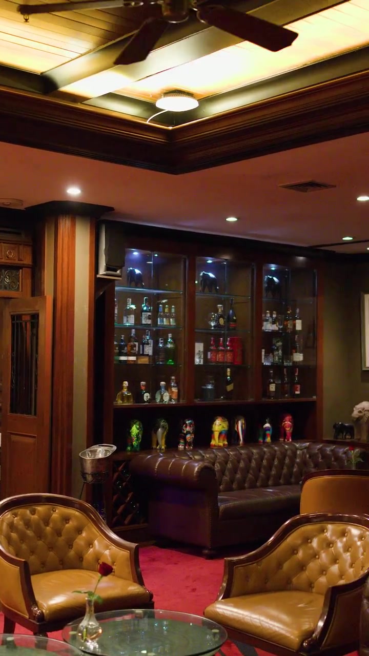 Lussuoso lounge bar con sedute in pelle, illuminazione calda, boiserie in legno e mensole espositive in vetro.