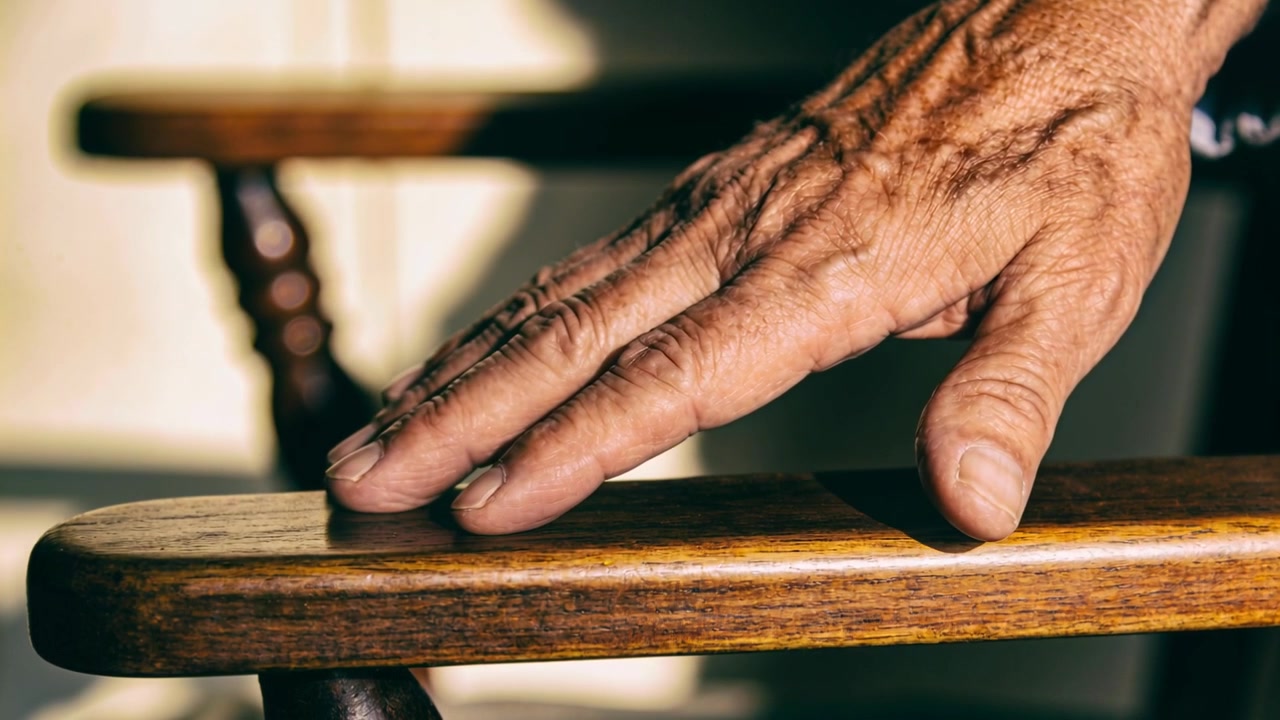 Mano anziana che si allunga e scivola lungo il bracciolo di legno a casa, cercando riposo con un polsino di colore chiaro.