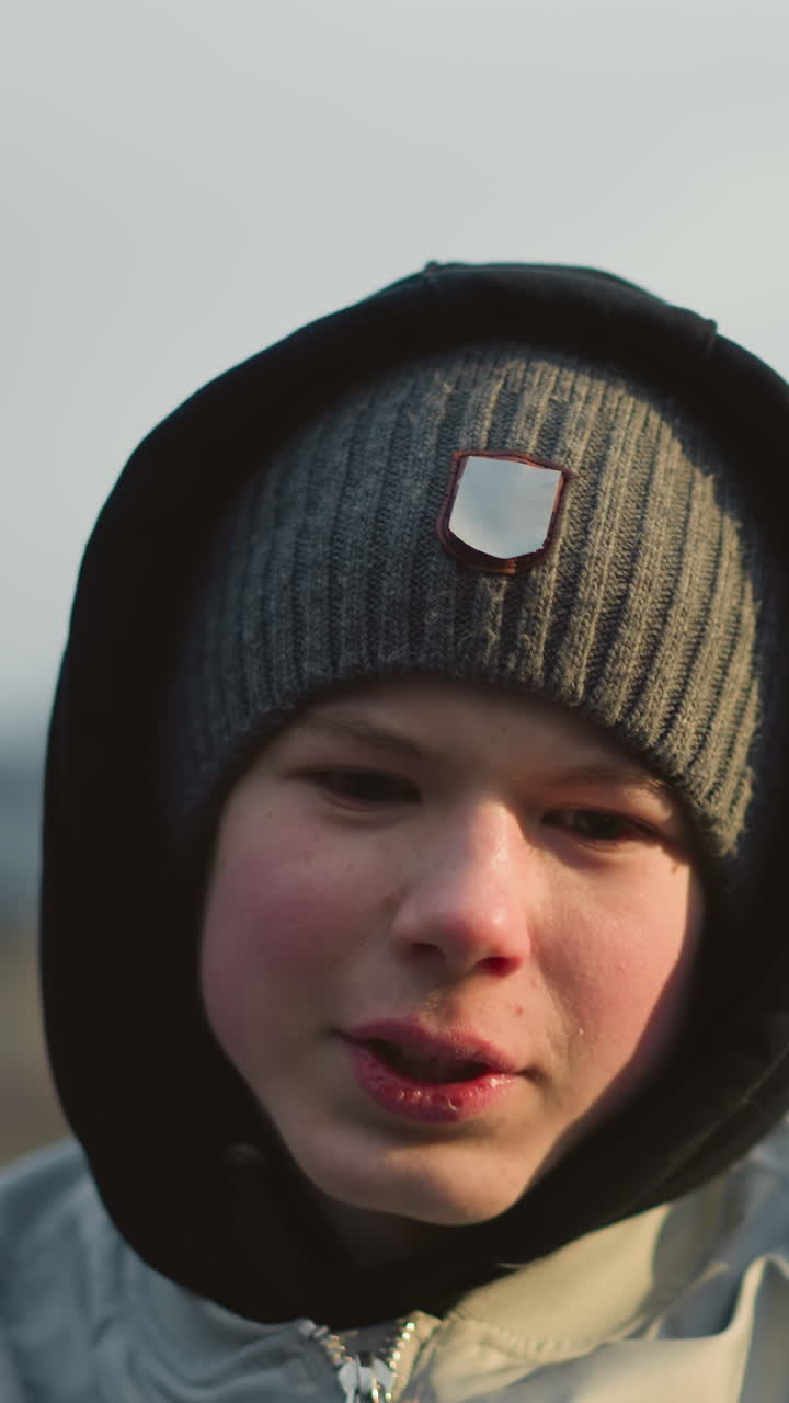 Close-up of a young boy wearing a gray top, beanie and a black hoodie, engaged in a discussion with someone, with a blur background of building