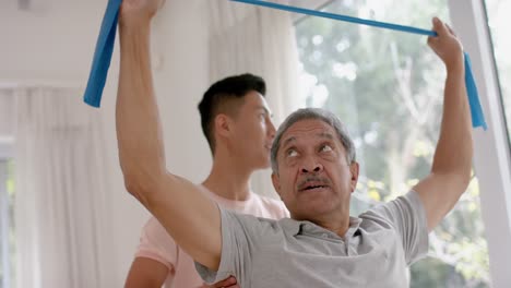 diverse male physiotherapist advising and senior male patient using exercise band, in slow motion