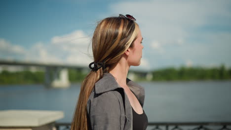 dama de camiseta gris y camiseta negra camina cerca de la orilla del río, pasando por una valla metálica, el fondo presenta una vista borrosa del puente y el agua mientras mira hacia el río y toma una respiración profunda