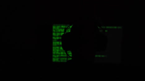 hacker sitting in front of a computer screen with green code in a dark shady room. blurred out screen.