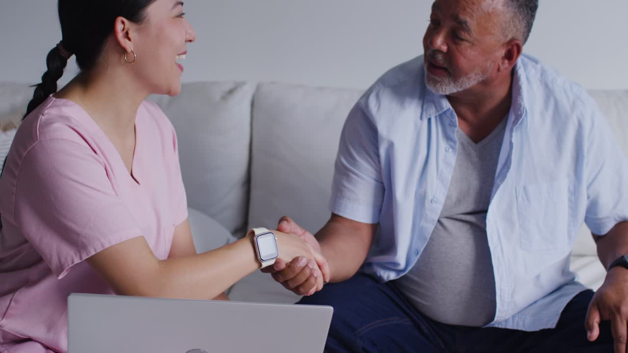 Donna asiatica in camice rosa che parla, protendendosi per una stretta di mano con un cliente uomo sul divano per offrirgli supporto.