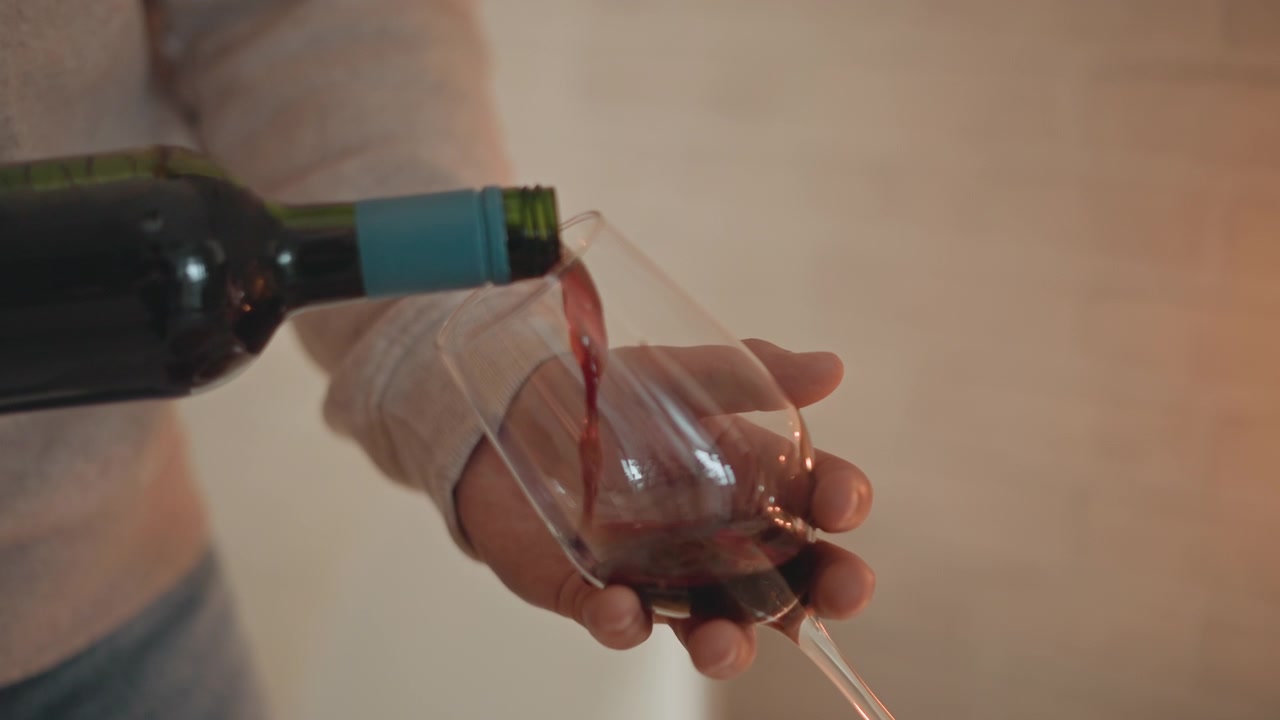 Des mains stables, vêtues d'un pull beige, inclinent une bouteille sombre tandis que du vin rouge coule dans un verre à pied sous une lumière intérieure chaude, montrant un versement contrôlé, des reflets doux et une atmosphère de repas détendue.