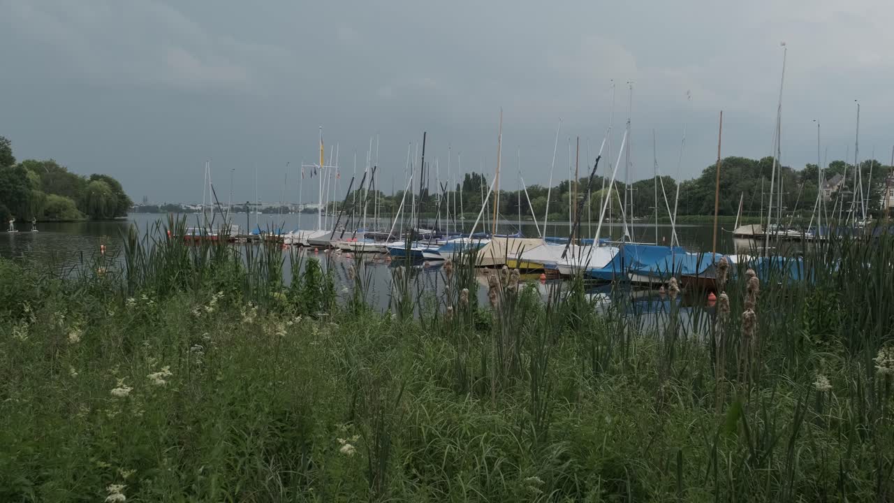 Streak of forked lightning seen over the city of Hamburg shrouded in grey cloud as seen from the Outer Alster lake with moored boats at a jetty over greenery in the foreground