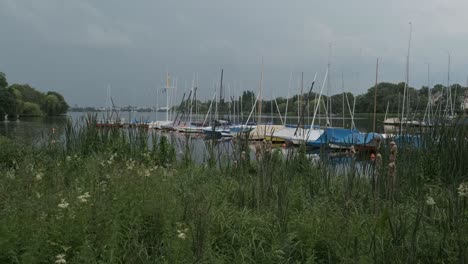 Streak-of-forked-lightning-seen-over-the-city-of-Hamburg-shrouded-in-grey-cloud-as-seen-from-the-Outer-Alster-lake-with-moored-boats-at-a-jetty-over-greenery-in-the-foreground