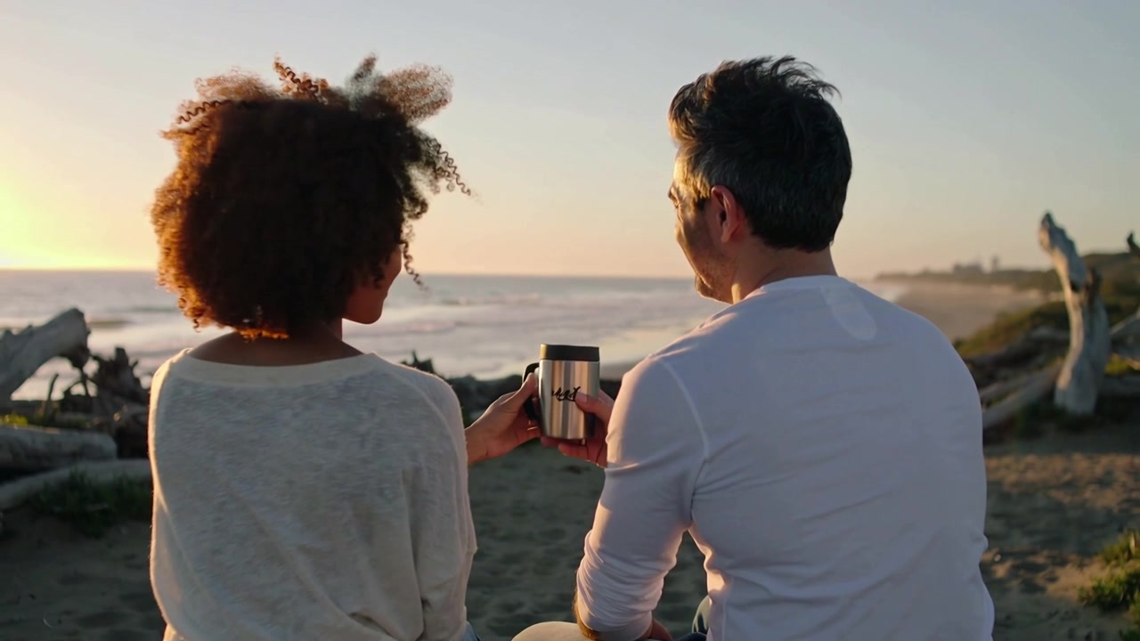 Offering man handing travel mug to woman wearing jeans on log, sharing drink as sun lowering