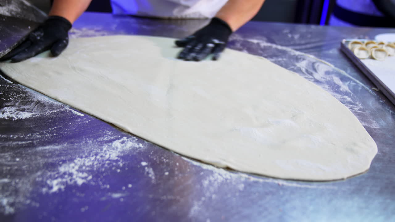 Cook's gloved hand spreads flour over the metal table. Rolled out white dough lies on the table for future cooking. Close up.
