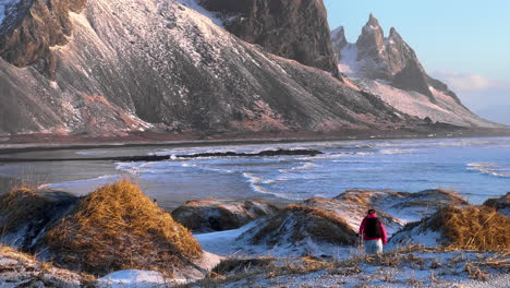 tourist hiking between snowy dunes and vestrahorn mountain in background during sunlight - stokksnes black sand beach