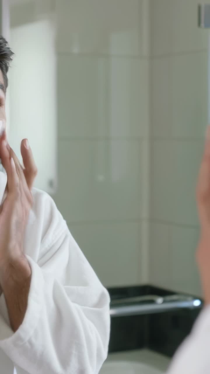 Vertical video: Facing mirror man applying cream across cheek for skin at home wearing white robe