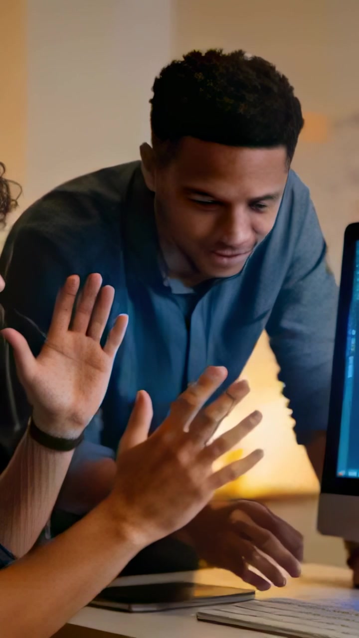 Vertical video: Leaning man blue top typing to edit at desk as peer pointing at screen with tablet