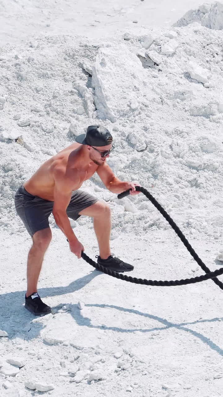 Sports activities with ropes on the street. Male athlete on the white sand does exercises with a rope. Vertical video