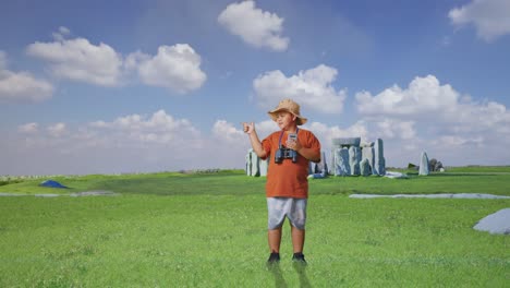 asian boy with a hat and binoculars looking at smartphone then smiling and pointing to side while traveling in stonehenge. boy researcher examines something, travel adventure, full body