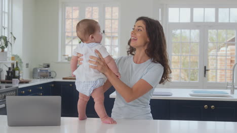 Mother-Playing-With-Baby-Son-Whilst-Working-From-Home-On-Laptop-In-Kitchen