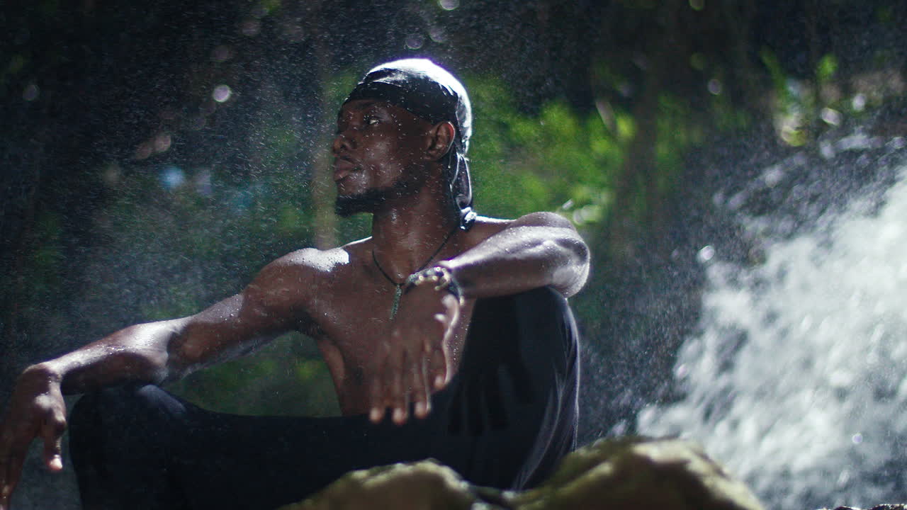 el joven modelo afroamericano se sienta quieto, mojado en la cascada de la selva tropical, el torso desnudo, respira tranquilamente, el agua fluye en el bosque natural, usa pañuelo, colgante y reloj