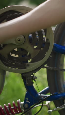 vista parcial de primer plano de la mano de alguien girando la rueda de una bicicleta, centrándose en la cadena y el sistema de engranajes, el fondo presenta una vista suavemente borrosa de la vegetación