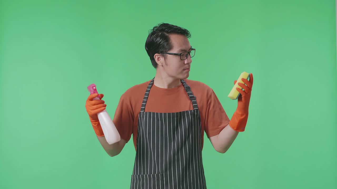 ama de llaves masculina asiática con un delantal mirando spray y esponja de cocina en las manos y luego sacudiendo la cabeza en el estudio de pantalla verde
