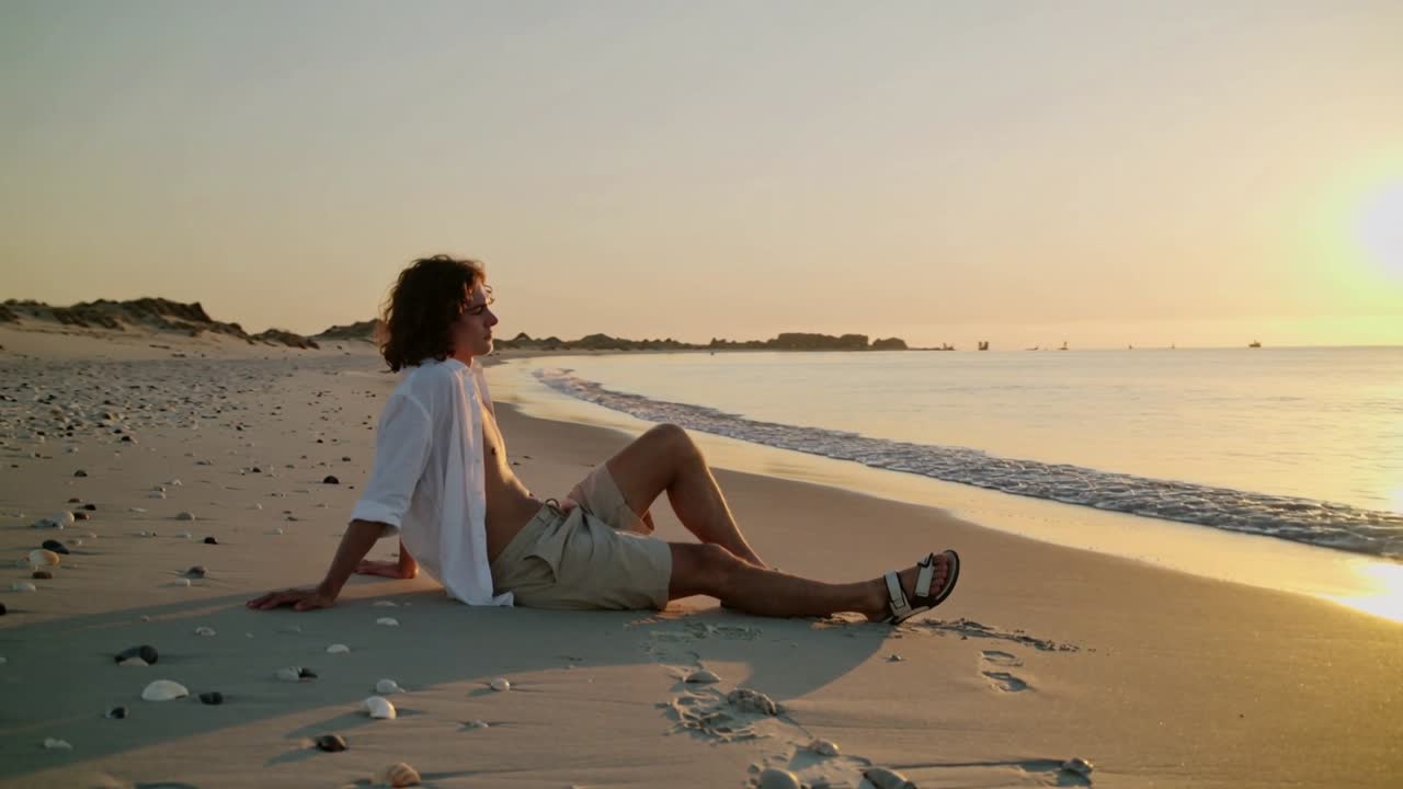 Sol poniéndose, hombre reclinado observando el horizonte en una playa de arena, con camisa abierta, gafas de sol y pantalones cortos.