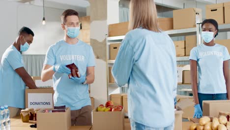 multiethnic group of volunteers in facial mask packing boxes with food and clothes and giving it to homeless people in charity warehouse