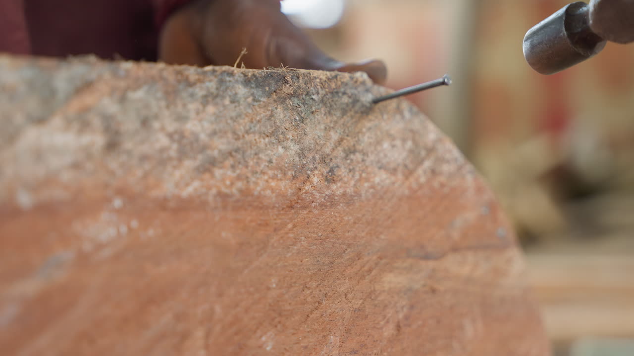 Close-up de mãos negras martelando um prego em uma tábua, alinhando o fixador de metal com golpes firmes, marceneiro concentrado fixando a tábua em uma bancada ensolarada, poeira e movimento rítmico transmitem a expertise do ofício.