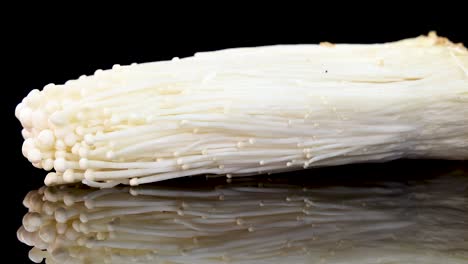a fresh enoki mushroom cluster slowly rotates on a glossy black background, with dramatic studio lighting and clear reflections enhancing its delicate texture