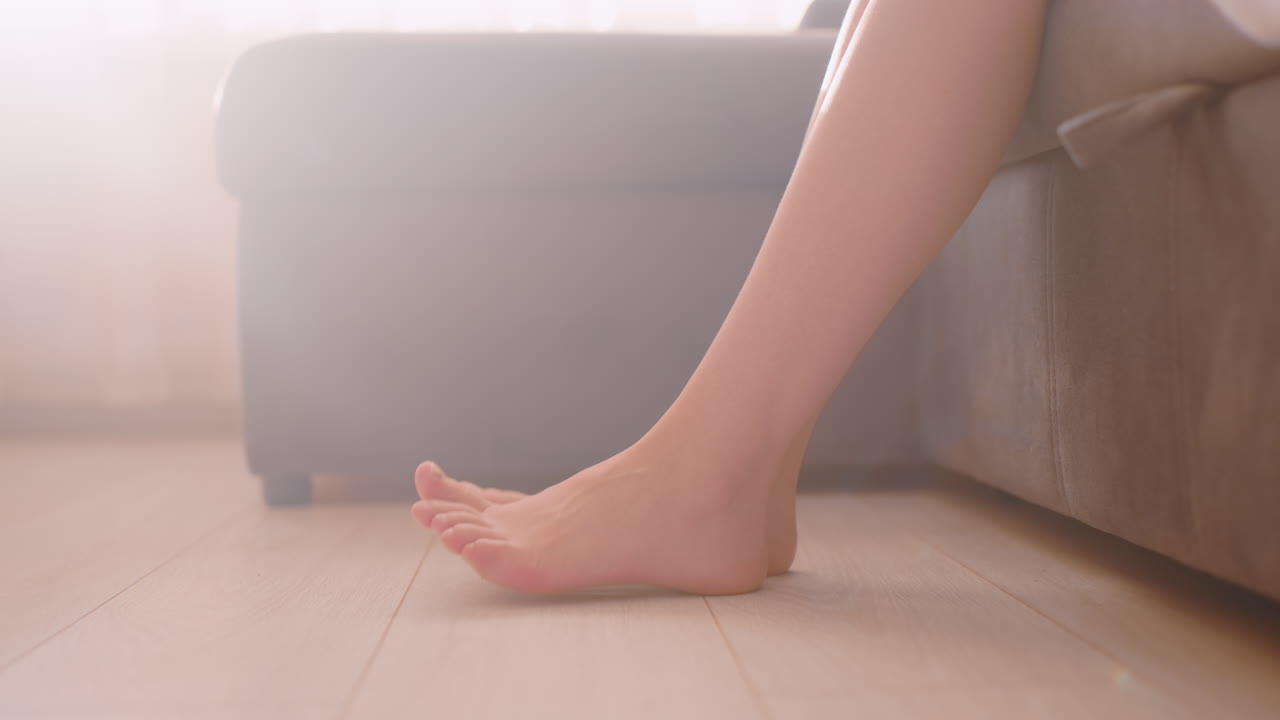 Side view of barefoot woman seated on soft couch lightly tapping toes on tiled wooden floor, surrounded by warm sunlight filtering through curtain, capturing soft relaxed indoor morning mood