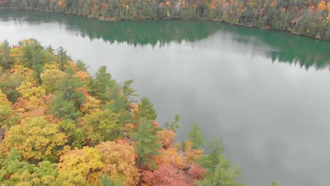 Rising-pan-to-the-left-aerial-shot-over-an-autumn-forest-beside-a-lake-with-the-trees-and-hills-reflection-in-the-blue-water-before-a-city-skyline-in-the-distance