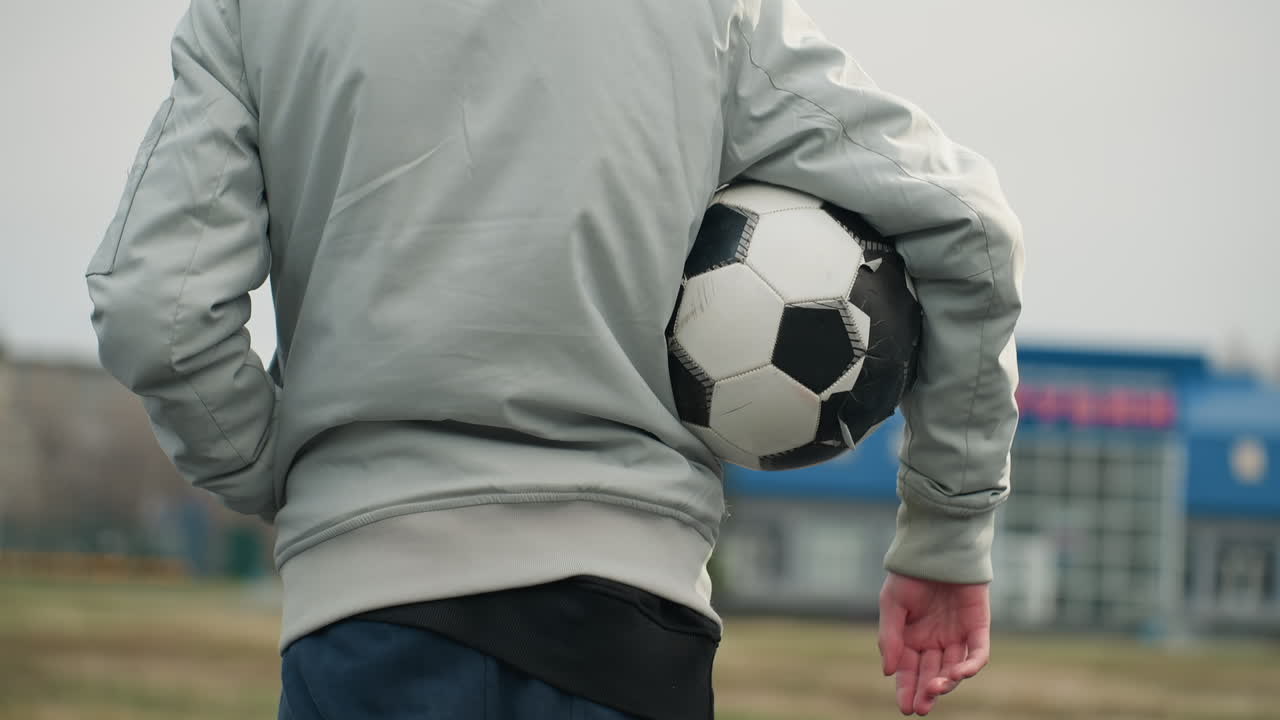 Back view of a person standing with a soccer ball tucked under their right arm, wearing a gray jacket and blue pants, with a blurred view of a grassy field and a blue building