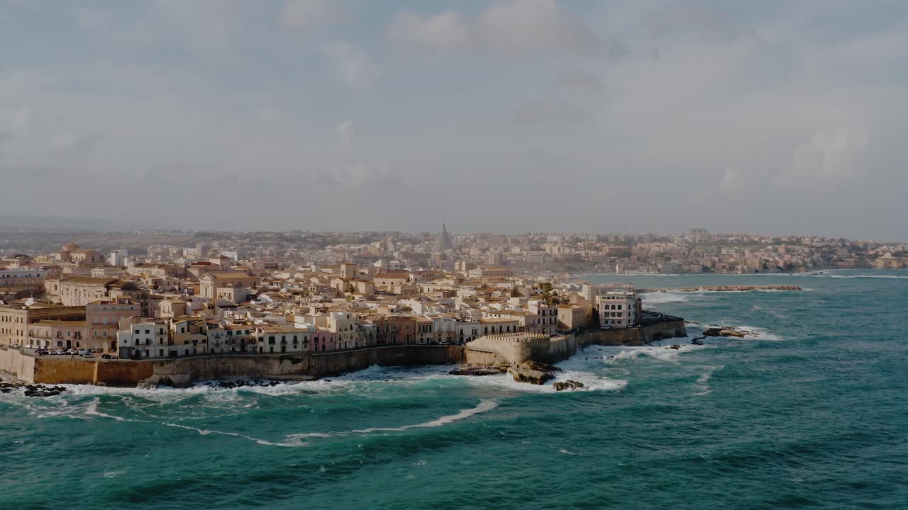 Wide panoramic aerial drone shot of Ortigia island. Historic architecture on a natural fortress by Mediterranean sea with large waves crashing on the coast. UNESCO world heritage site in Sicily.