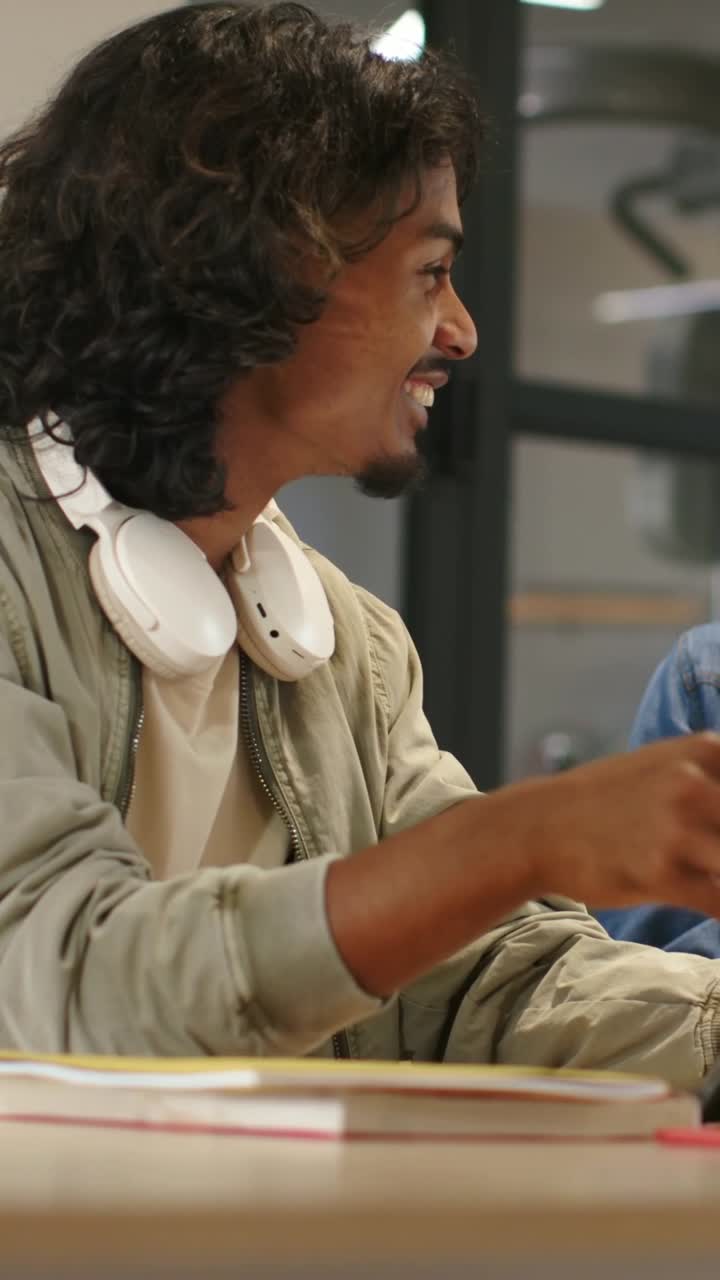 Vertical video: Reaching man in olive coat as peer showing phone at desk with notes and headphones