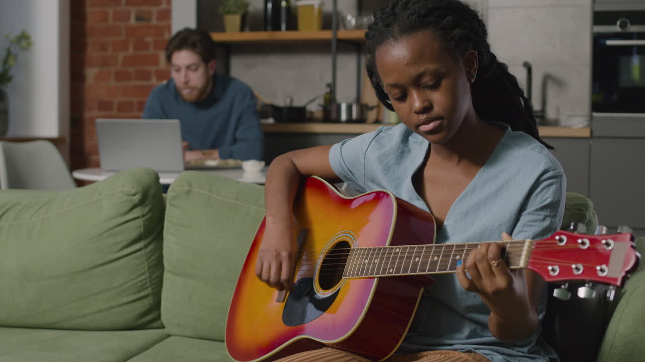Girl Playing Guitar Sitting On Sofa While Her Male Roommate Using The Computer 1