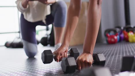 Video-of-diverse-female-fitness-trainer-and-determined-woman-raising-dumbbells-working-out-at-a-gym