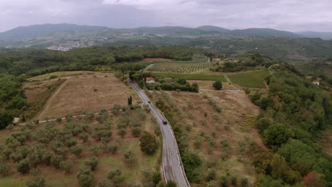 Drone-catching-up-with-a-black-car-driving-over-a-asphalt-road,-surrounded-by-nature-environment