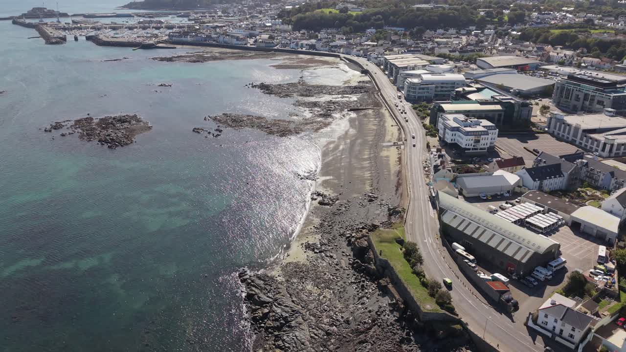 High drone footage of Belle Greve Bay Guernsey heading south along Les Banques and Admiral Park at mid tide in bright afternoon sun with clear sea and rocks and views to St Peter Port