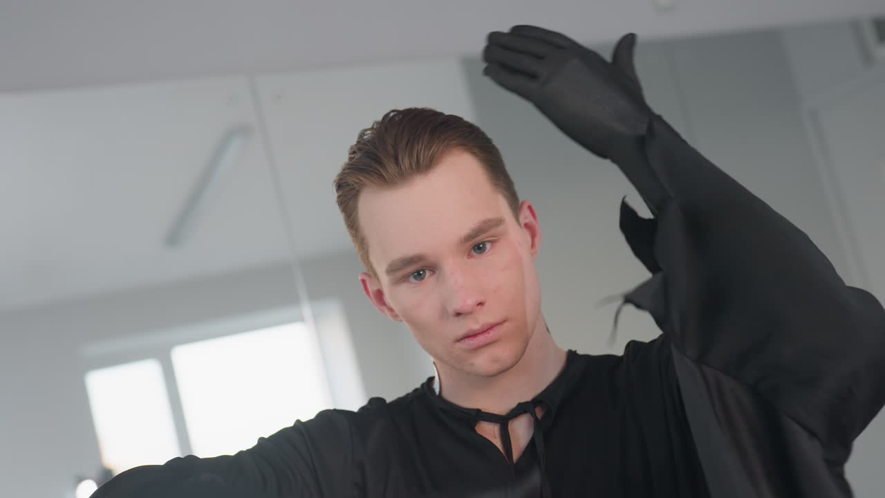 young boy in dark theatrical costume raises arm gracefully in front of mirror with confident facial expression and slight smile, performing subtle movement gesture in bright indoor setting