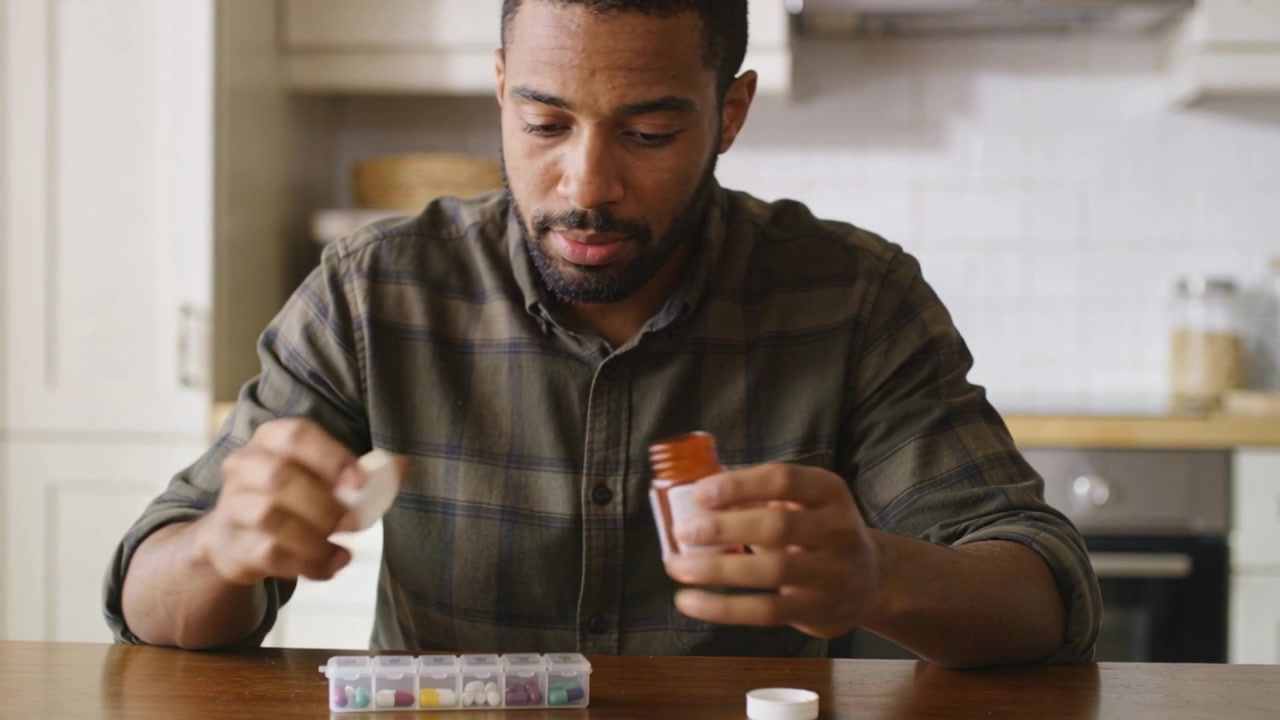Filling man plaid-shirt pouring meds from amber bottle into weekly box at kitchen table to organize