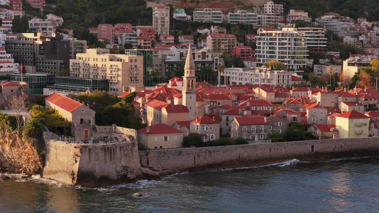 Vista aerea da drone della città vecchia di Budva, che mostra i suoi edifici medievali in pietra, i tetti rossi, le mura della fortezza e il campanile della chiesa lungo la costa, con edifici moderni e montagne sullo sfondo. Montenegro