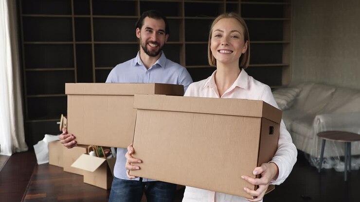 Happy couple standing in living room holding boxes with belongings