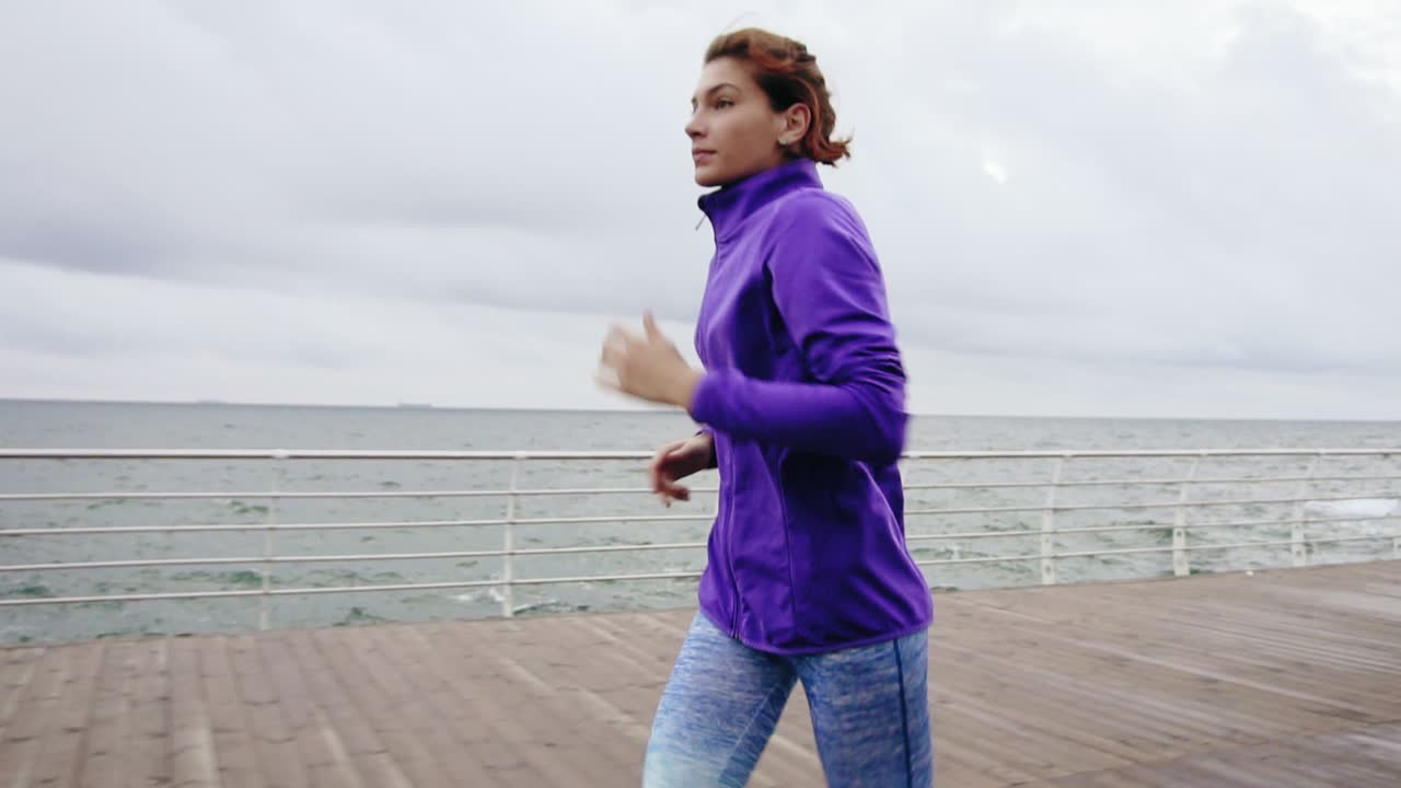 Primo piano di giovane donna atletica che corre all'aperto al rallentatore su una passeggiata vicino all'oceano, godendosi la corsa mattutina.
