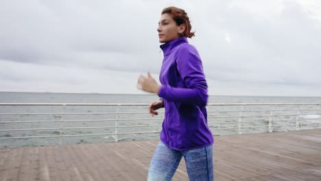 Vista-Cercana-De-Una-Joven-Atlética-Corriendo-Al-Aire-Libre-En-Cámara-Lenta-En-El-Paseo-Marítimo-Cerca-Del-Océano-Disfrutando-De-Una-Carrera-Matutina