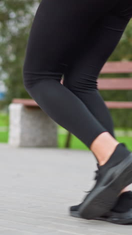 close-up view of woman's legs in black leggings and sneakers running in park path, showing motion, fitness routine, and outdoor jogging with park bench and nature in background