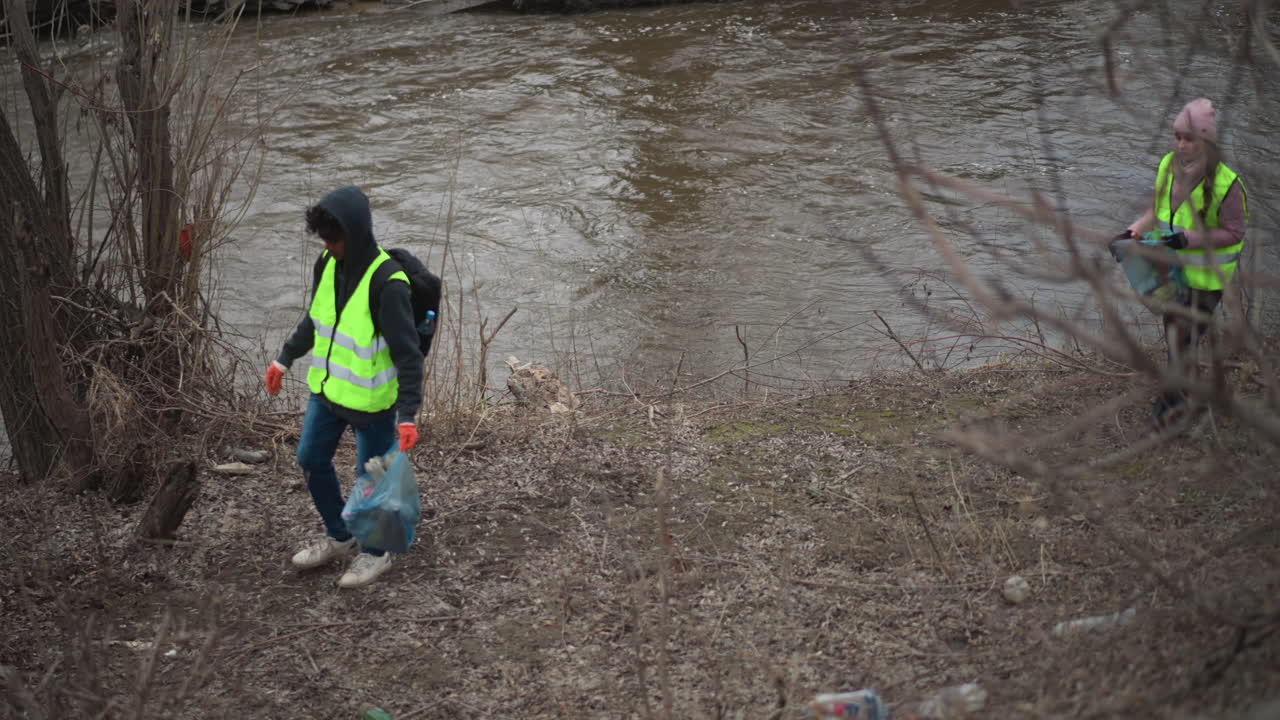 Volunteer in reflective vest with gloves and backpack picking up plastic waste and trash along polluted riverbank on cold overcast day, surrounded by leafless trees during environmental cleanup effort